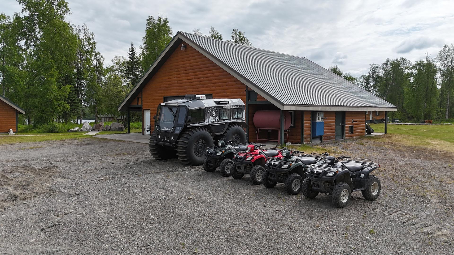 Log cabin-style building with a large black monster truck and a row of ATVs parked in front on a gravel lot, forest in the background.