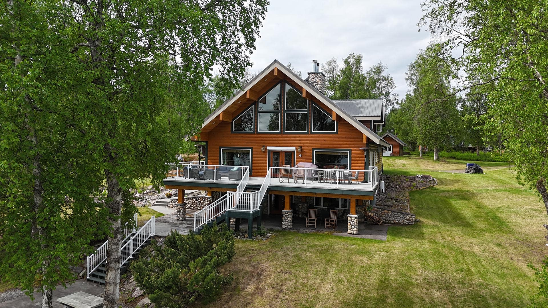 Log cabin with a large gable of glass windows and a wraparound deck amid green trees.