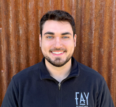 Smiling man in a black zip-up fleece with a FAY RANCHES logo, standing in front of a rusted corrugated metal wall.