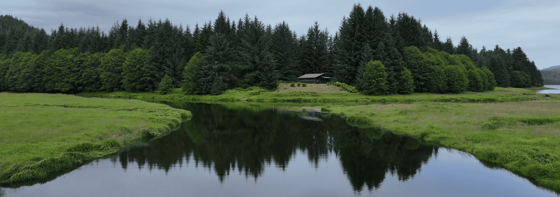 A red cabin sits among evergreen trees beside a winding river, with lush green fields in the foreground.