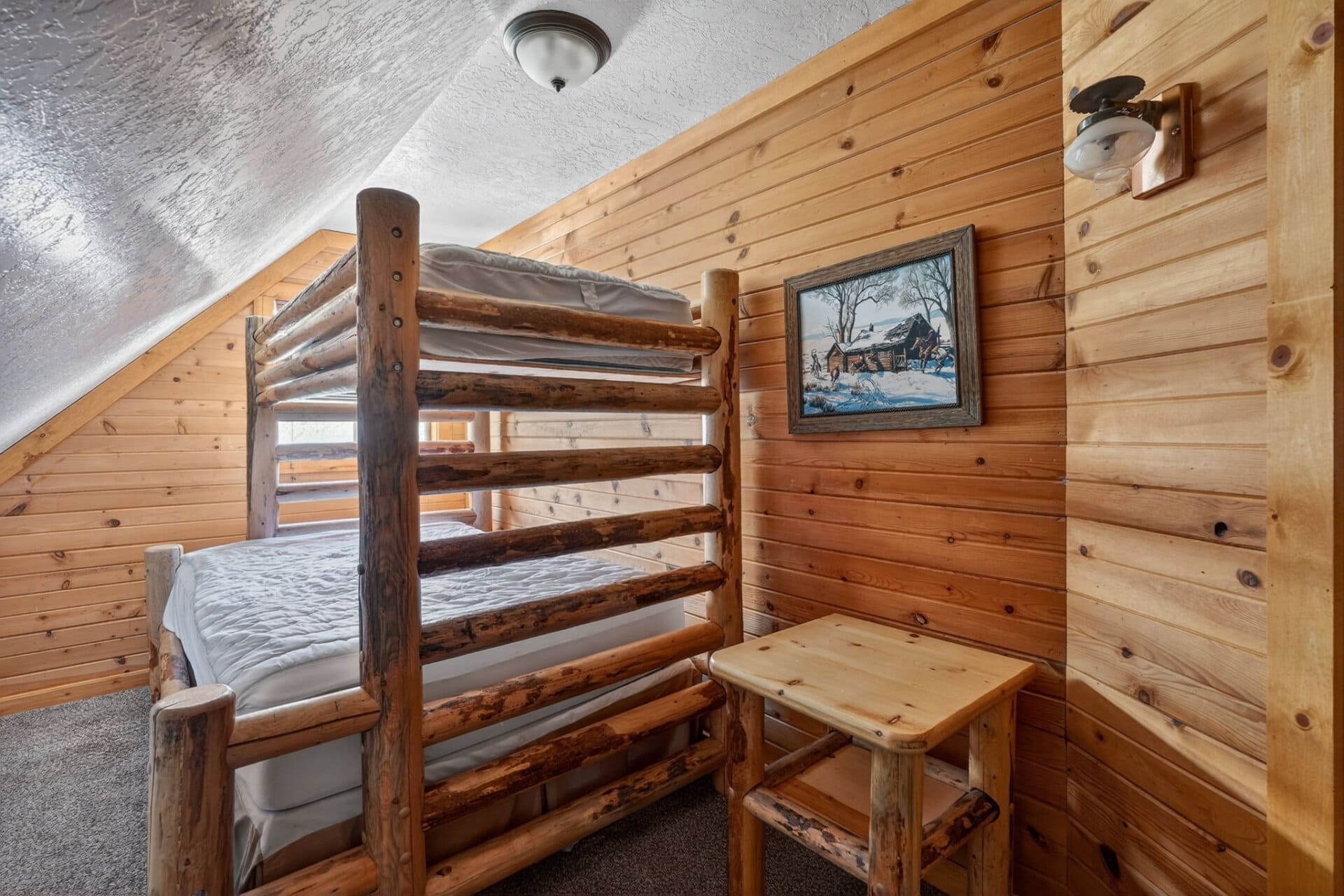 Rustic log bunk bed in a wood-paneled cabin room, with a framed winter landscape painting on the wall and a small wooden table nearby