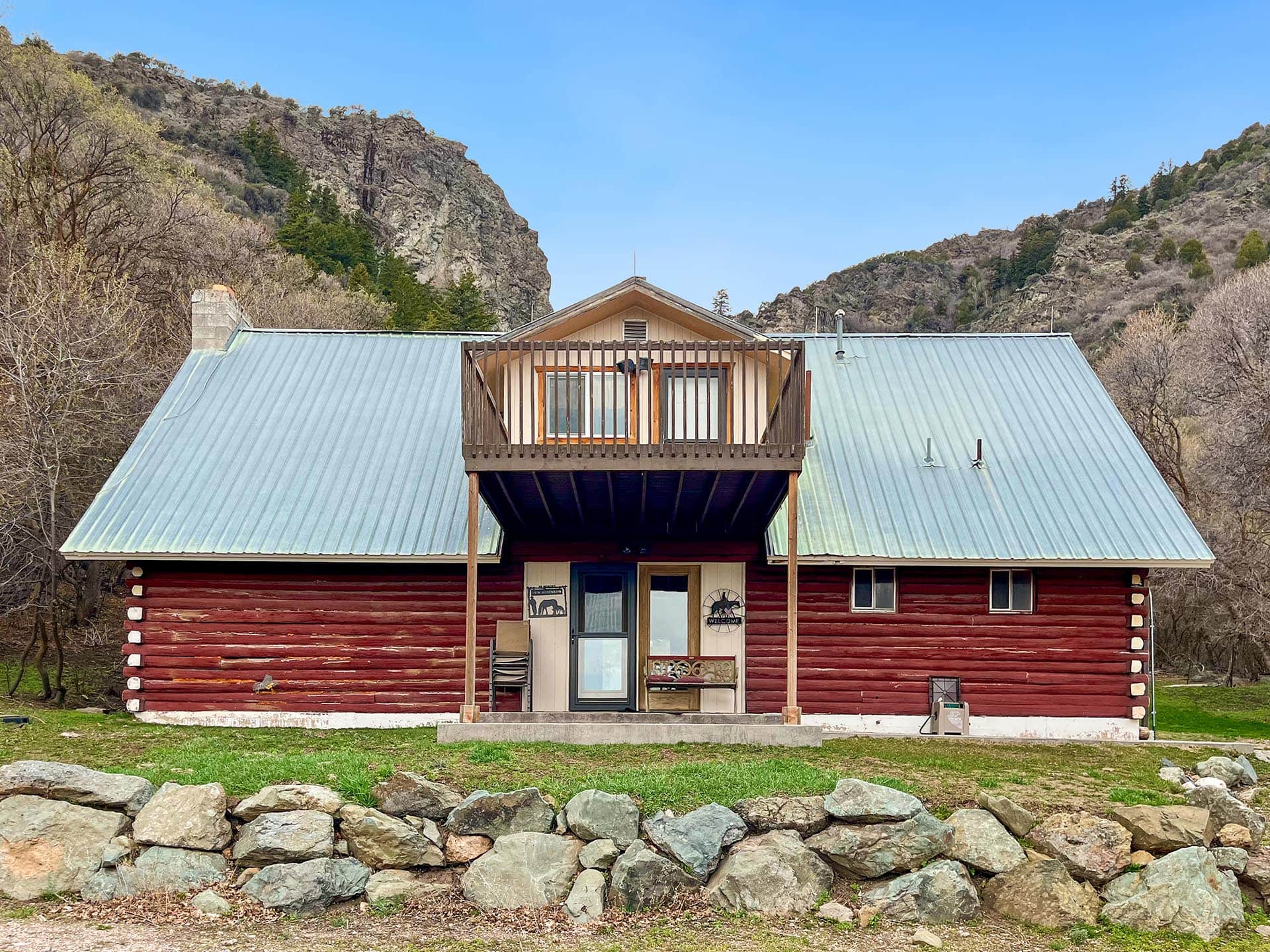 Red log cabin with a metal roof and small balcony, set against rocky hills and trees.