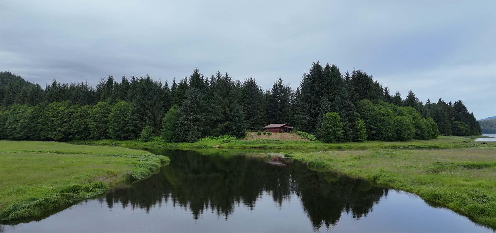 Small red cabin nestled in a dense pine forest, beside a calm reflective river and green meadows under an overcast sky.
