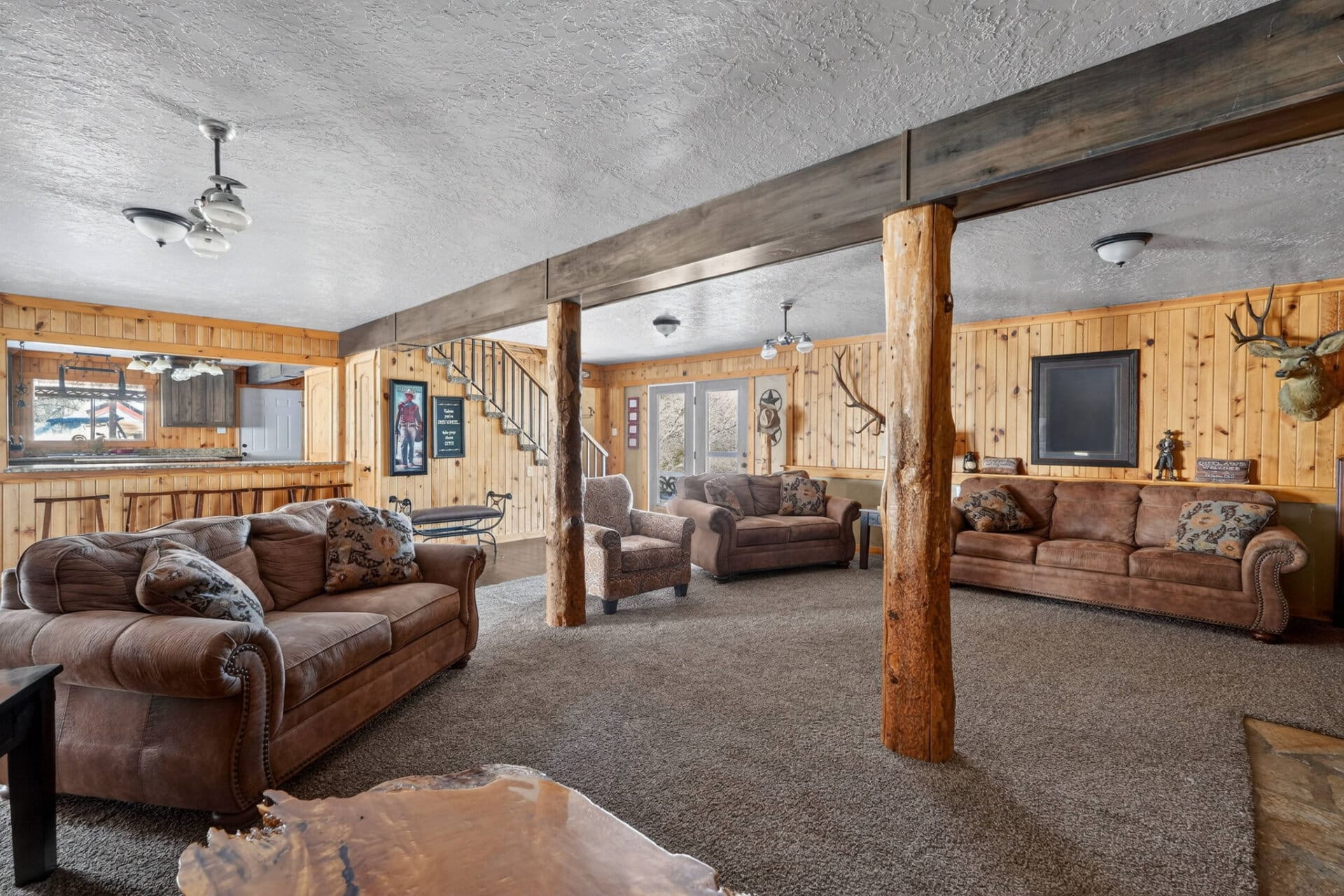 Rustic living room with brown leather sofas, wooden panel walls, and exposed log beams; deer head mounted on the wall.
