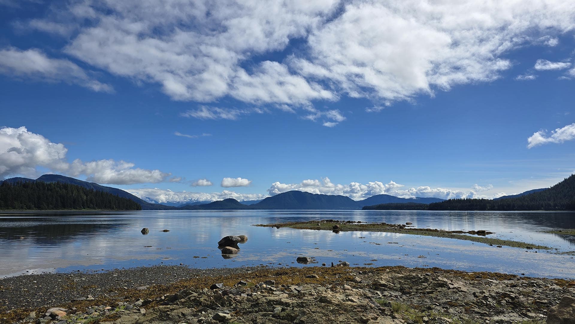 Tranquil alpine lake with forested mountains on either shore under a bright blue sky with scattered white clouds, Rocky shoreline in the foreground.