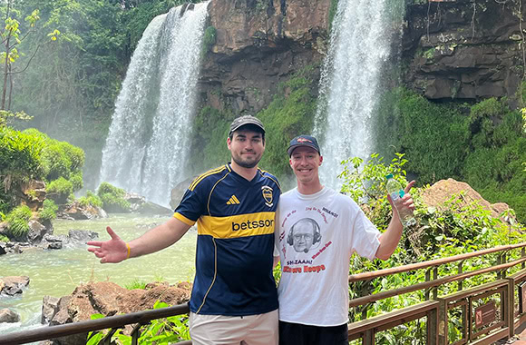 Two men pose with open arms in front of a tall waterfall, on a wooden railing amid lush greenery.