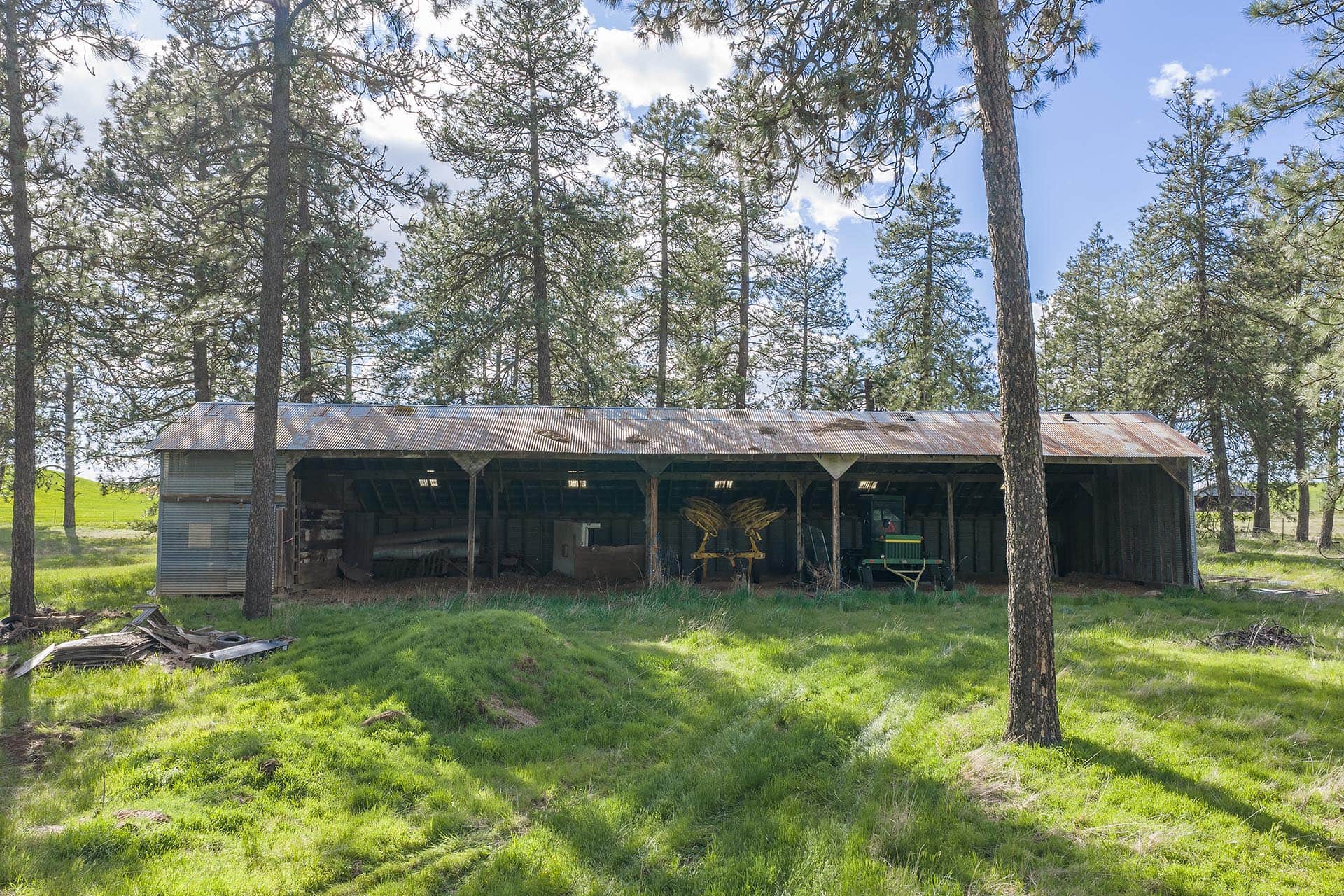 Metal-roof barn in a pine forest with farming equipment visible inside the open bays beneath the roof