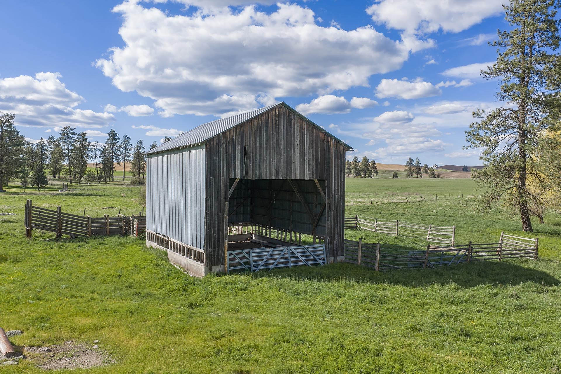 Old weathered wooden barn in a grassy rural field with fencing and trees under a blue sky with clouds