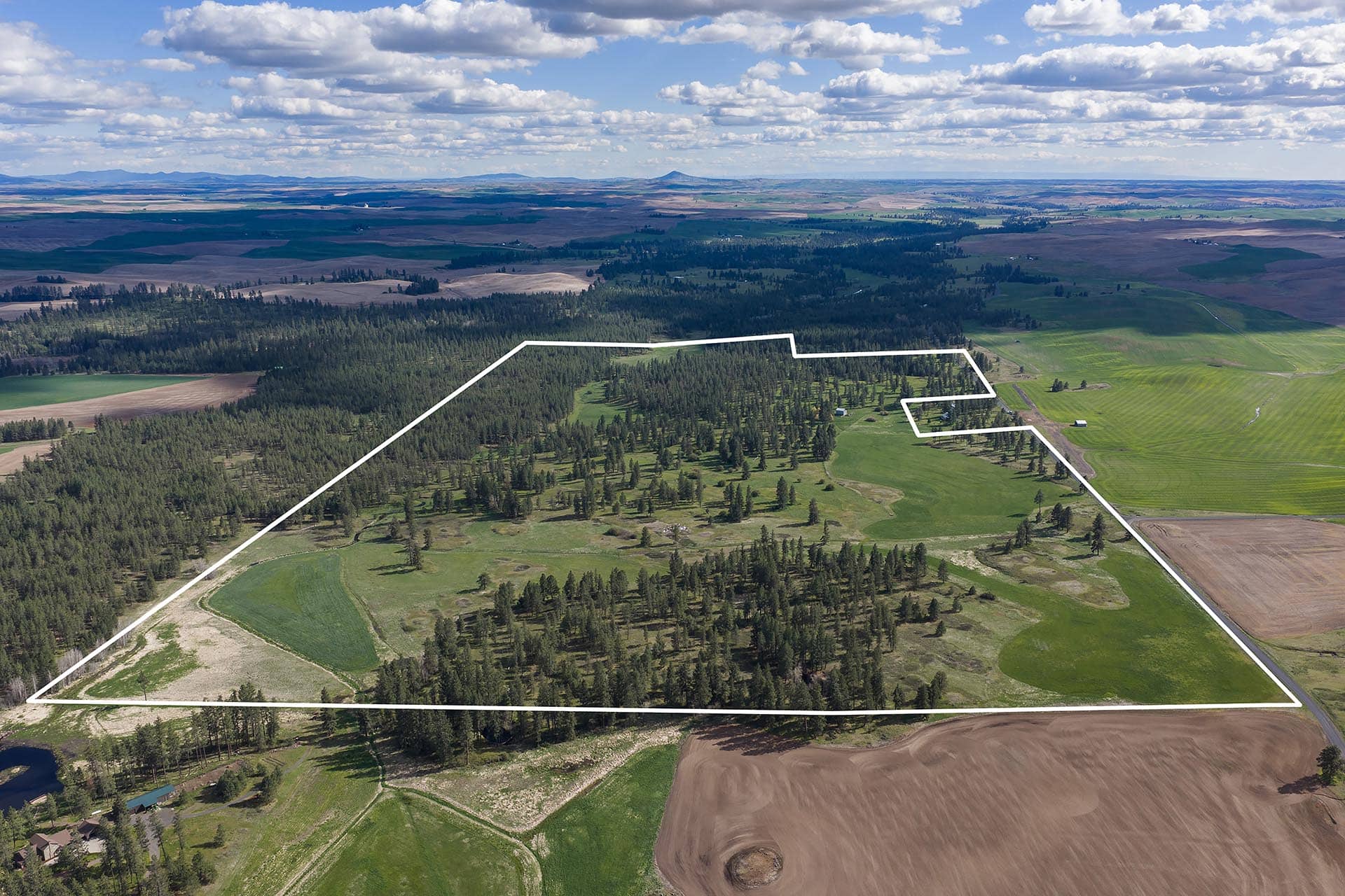 Aerial view of farmland with a white outline marking a large irregular property boundary, showing fields and forests spread across rolling terrain.
