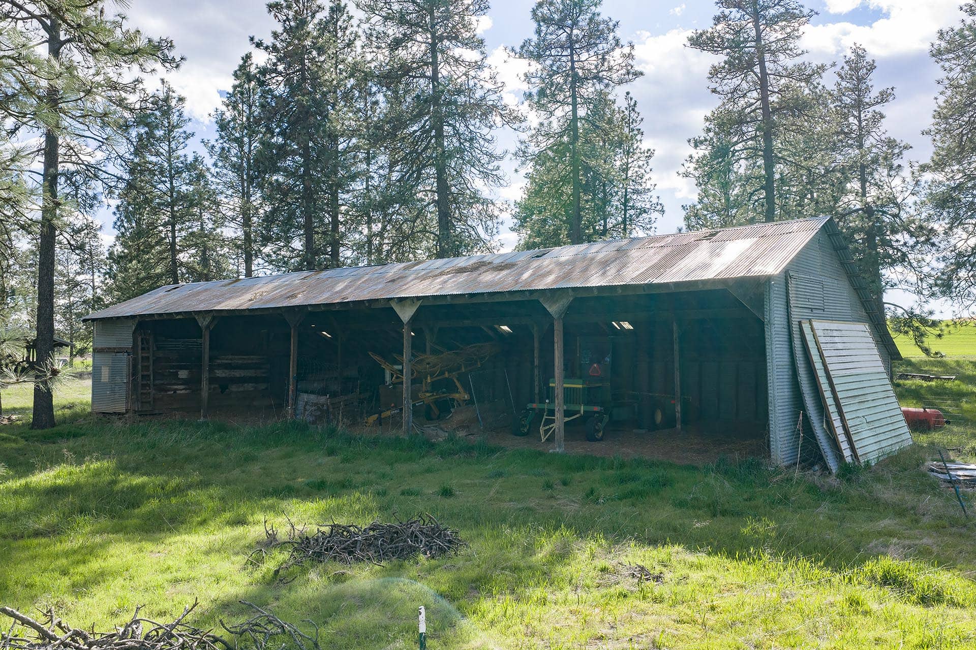 Open-sided metal-roof barn in a grassy field, surrounded by tall pine trees with farm equipment visible inside.