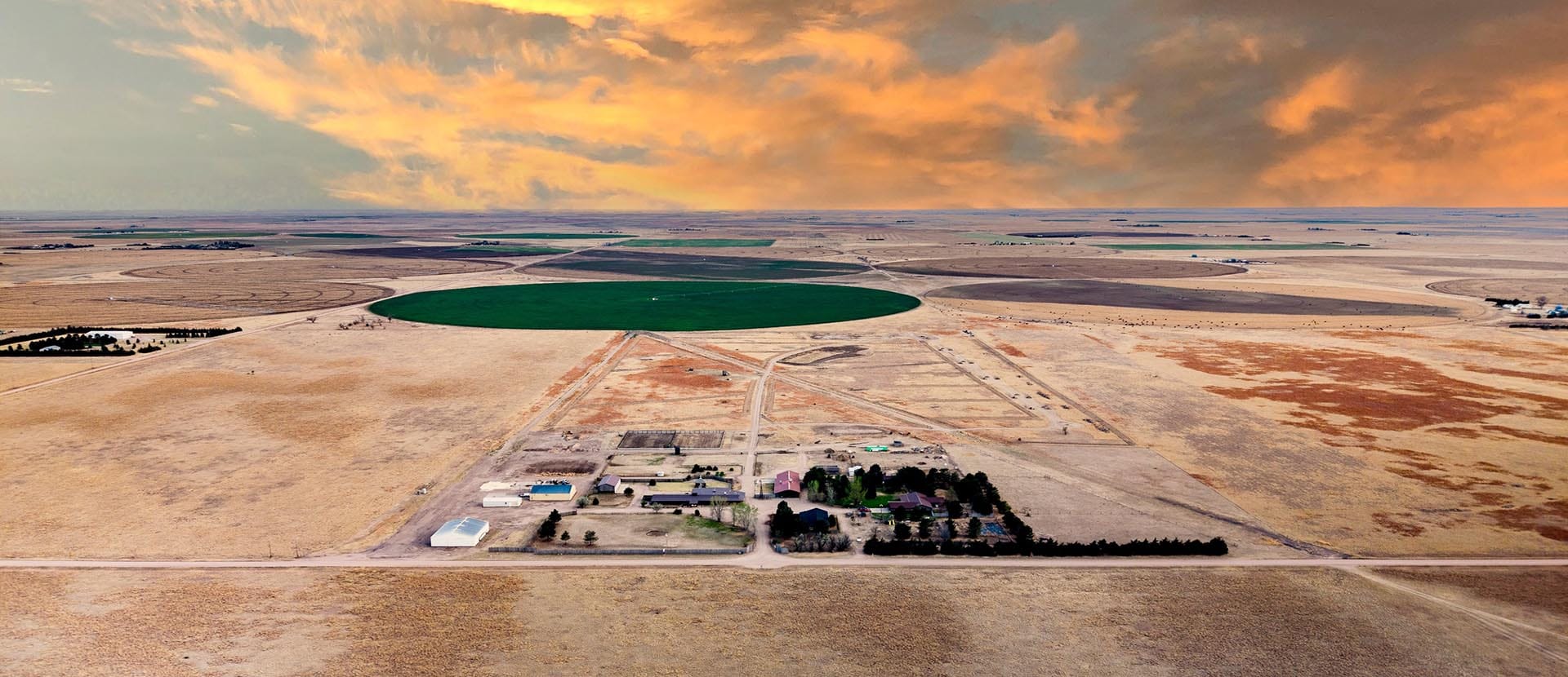 Aerial view of circular irrigation fields on a dry plain, with a small farm cluster at the bottom and an orange sunset sky.