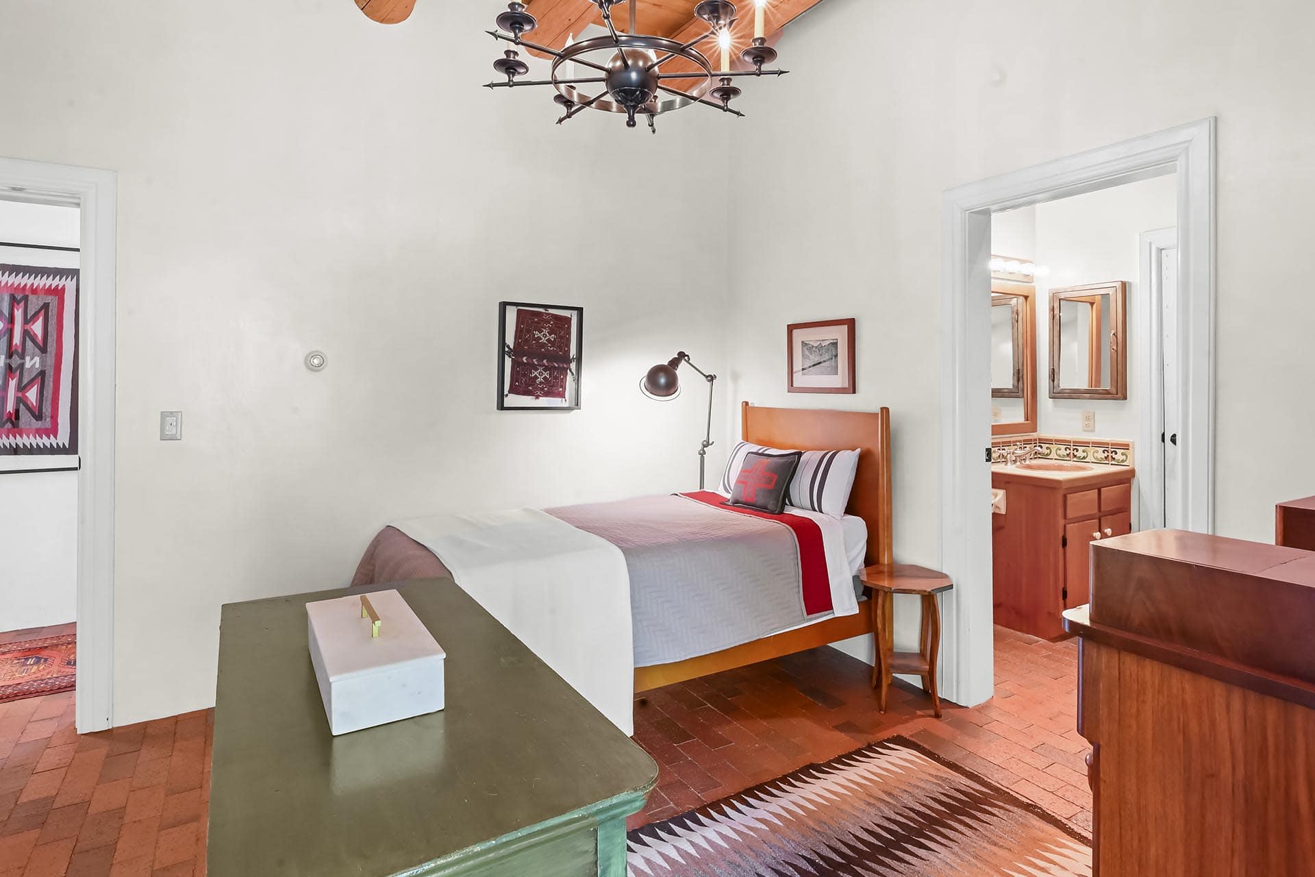 Bedroom with a wooden bed, gray-red quilt, and a green foot bench; doorway to a bathroom visible on the right.