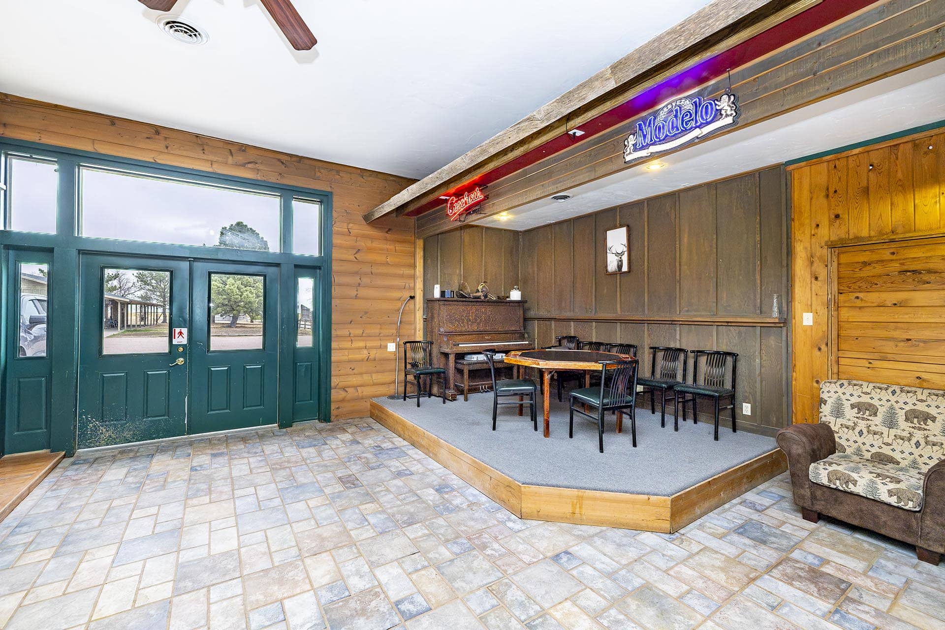 Indoor lounge with a wooden stage, piano, and several chairs beneath neon signs (Modelo, Coca-Cola) on a rustic wall. High glass green doors to the left.