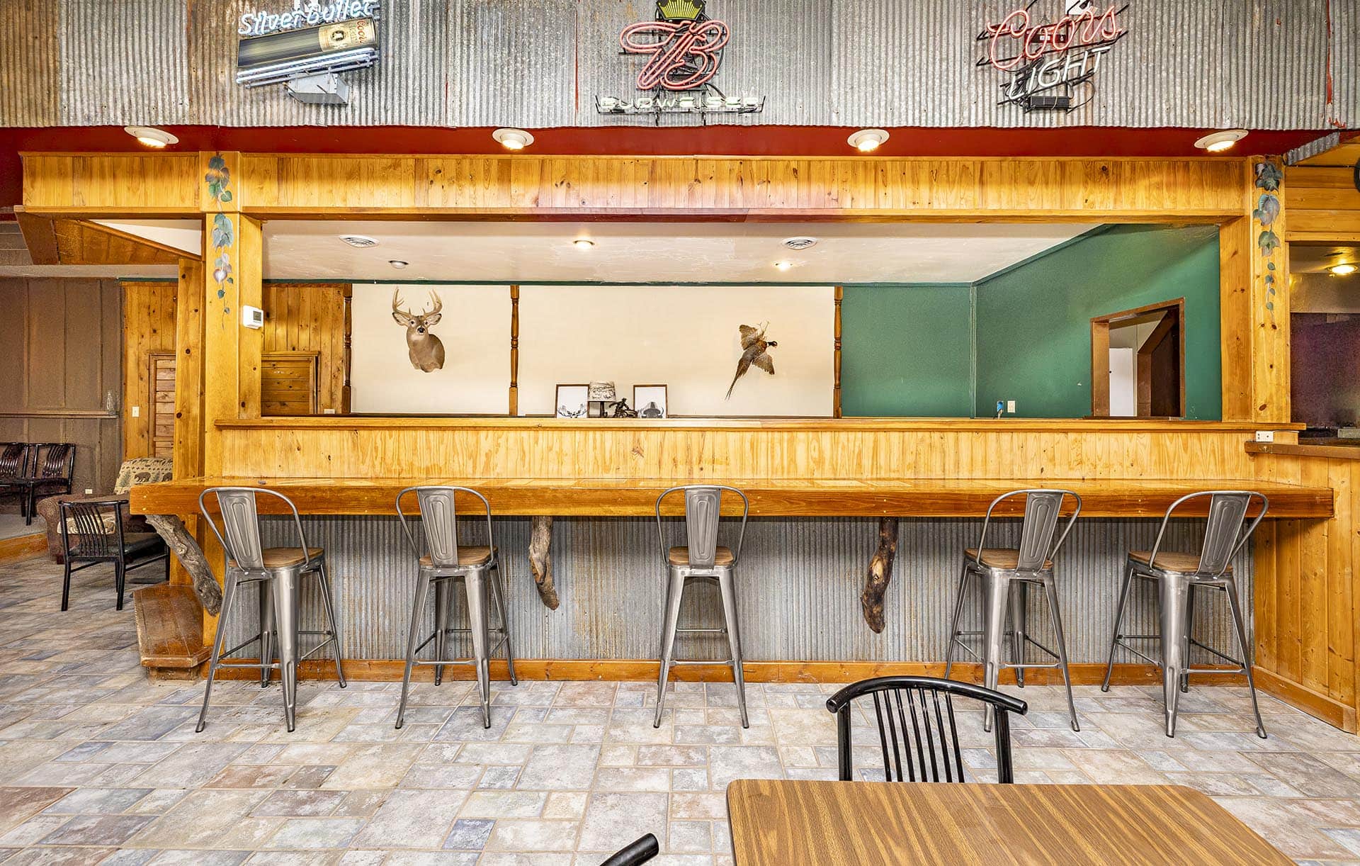 Rustic bar interior with a long wooden counter, metal stools, and deer head decor on the wall.