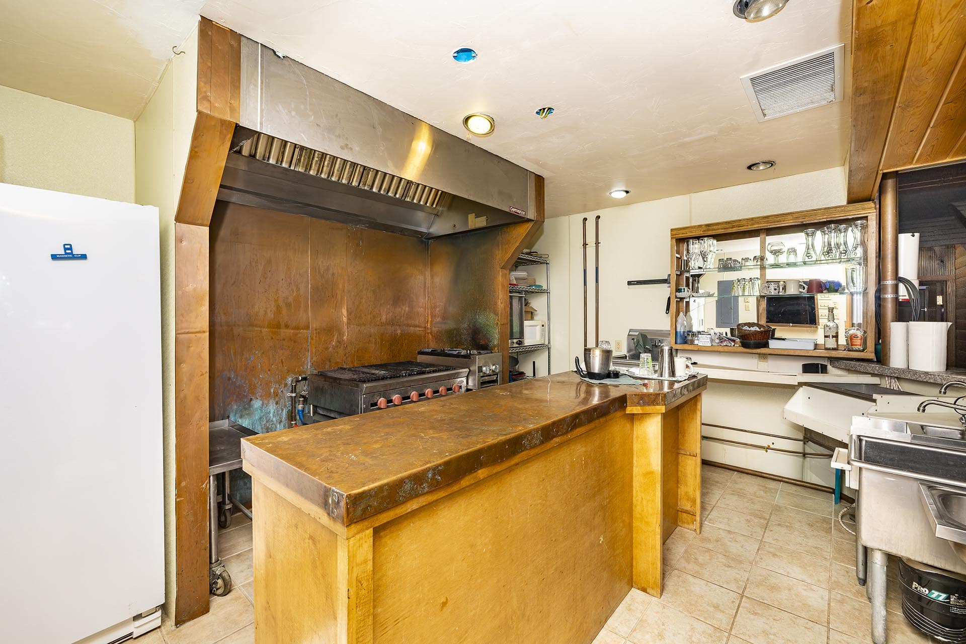 Industrial-style kitchen with a large stainless steel vent hood over a gas grill and a rust-colored metal wall behind it, framed by wooden supports and a wooden prep island in the foreground