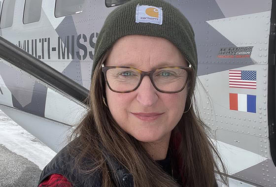 Portrait of a woman with glasses wearing a green Carhartt beanie, standing in front of a gray helicopter with U.S. and French flags visible.