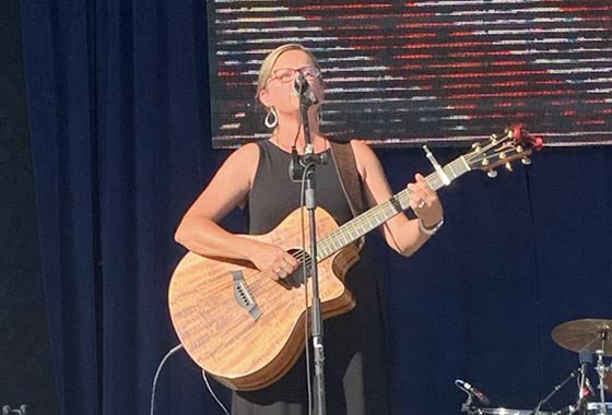 Female musician in a black dress sings into a microphone while playing an acoustic guitar on a stage with a dark blue backdrop and a large display behind her.