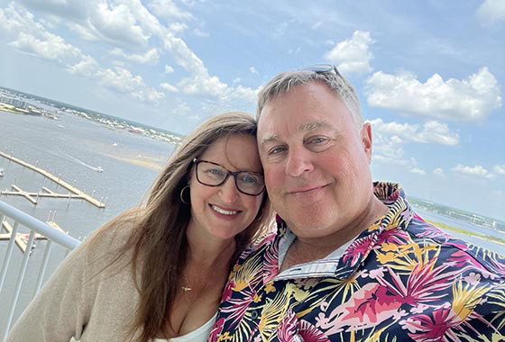 Smiling couple posing on a sunny boardwalk with the ocean and blue sky in the background.