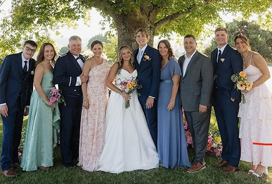 Bride and groom pose with their wedding party outdoors beneath a leafy tree; bridesmaids in pastel dresses and groomsmen in suits hold bouquets and smile on the lawn under flowers.