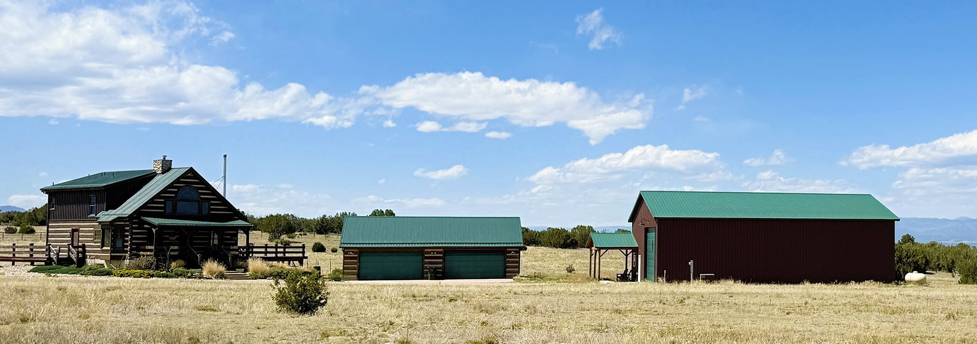 Rustic farmstead in a dry field: a log cabin with a green roof, two green-roof garages, and a large red barn under a bright blue sky.