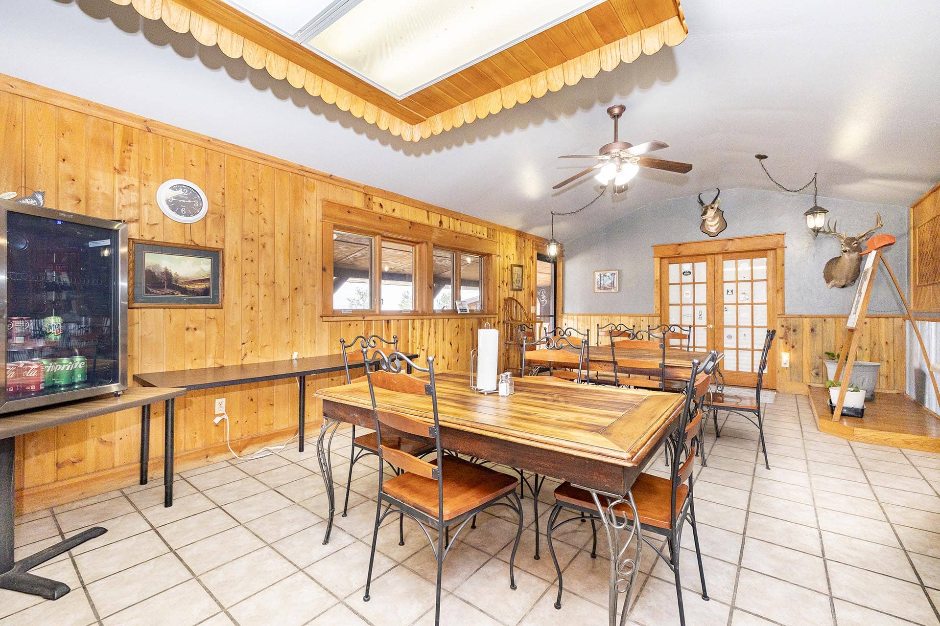 Rustic dining room with wood-paneled walls, long wooden tables, and metal chairs; hunting trophies on the blue wall above the doors.