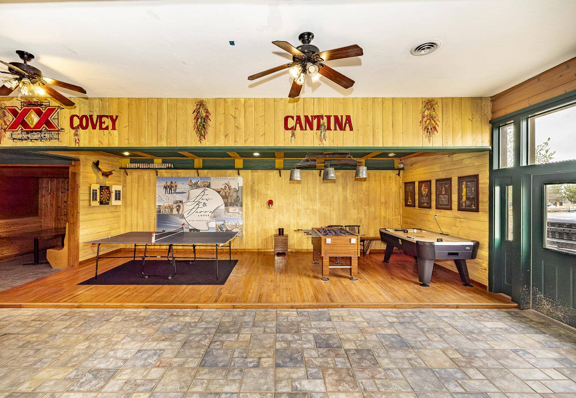 Cantina-style game room with wood-panel walls, ceiling fans, and tables for ping-pong, foosball, and pool under warm lighting.