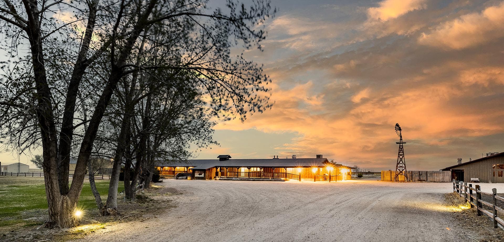 Dusk at a rural farm: gravel road leads to a lit wooden barn with a windmill on the right and trees lining the left edge of the scene