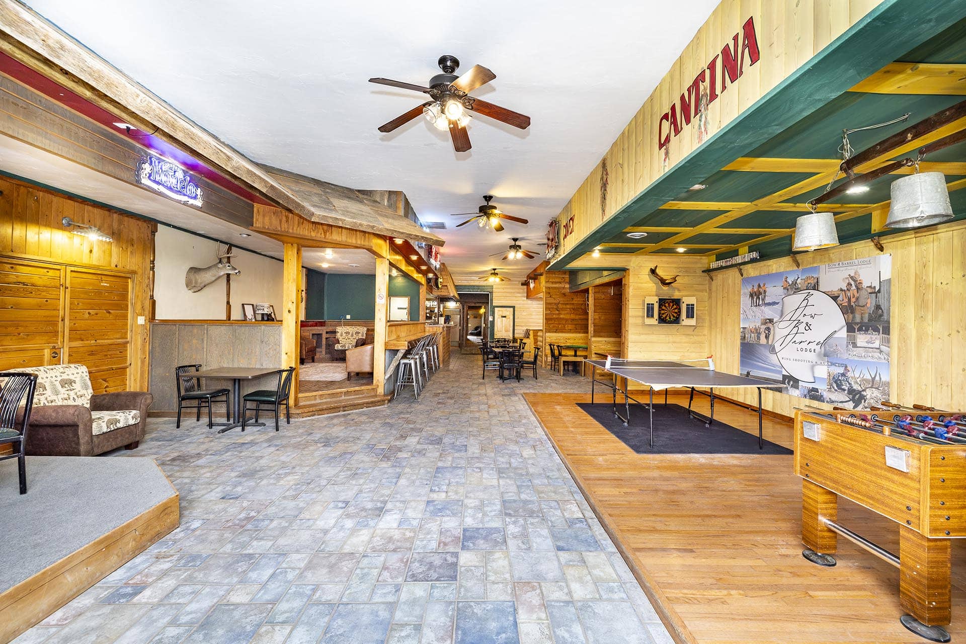 Interior of a rustic lodge lounge with wood paneling, ceiling fans, and mixed seating along a long hallway-like space.