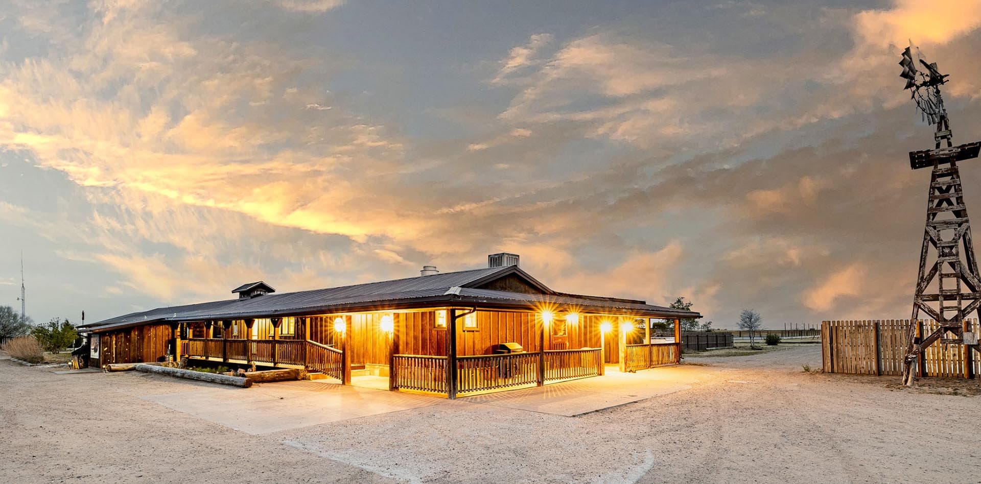 Wooden ranch building at dusk with warm lights, a wraparound porch, and a tall windmill on the right in a desert landscape.