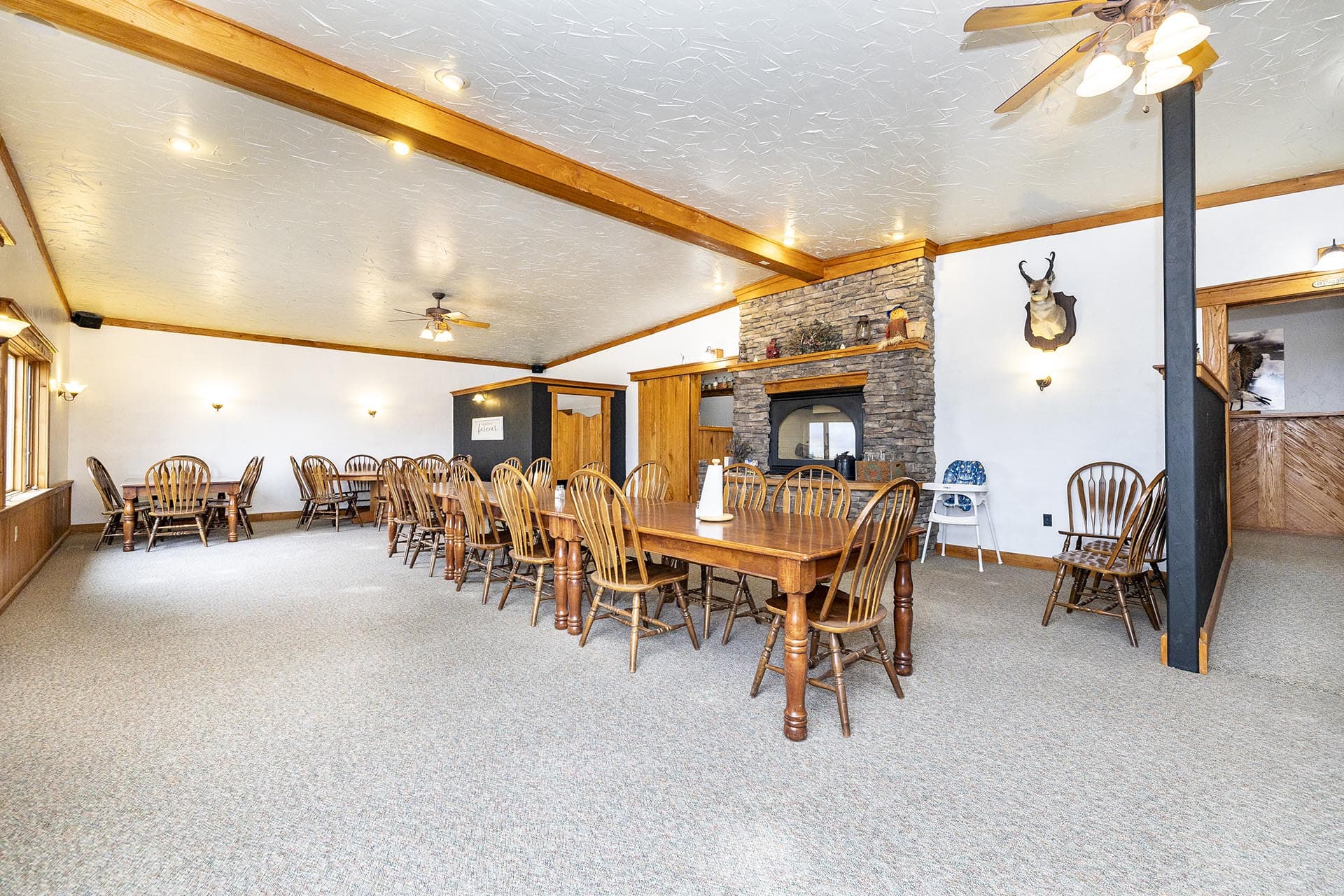 Rustic dining room with a long wooden table, many chairs, and a stone fireplace under wooden beams.