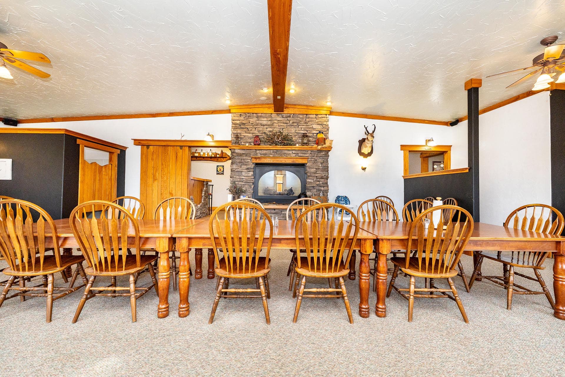 Rustic dining room with a long wooden table and numerous matching chairs facing a stone fireplace at the far wall.