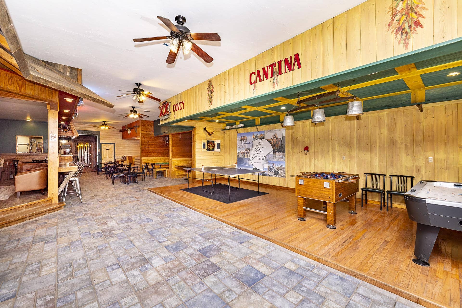 Rustic indoor game room with wood-paneled walls, ceiling fans, and a cantina sign; contains a ping-pong table, foosball, and lounge seating.