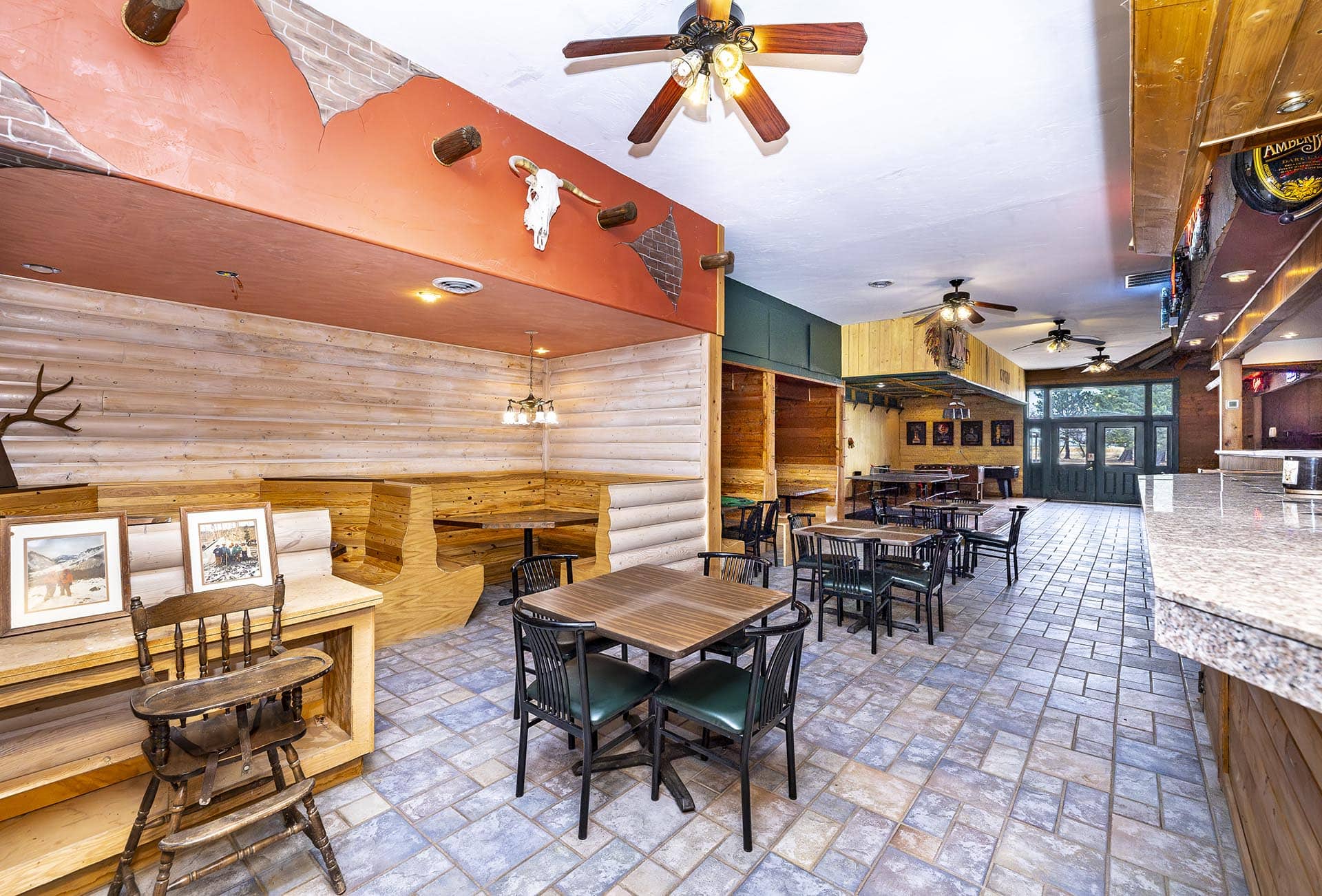 Rustic dining area with wooden walls, multiple tables and chairs, and a deer skull mounted on the orange wall above a wooden bench.