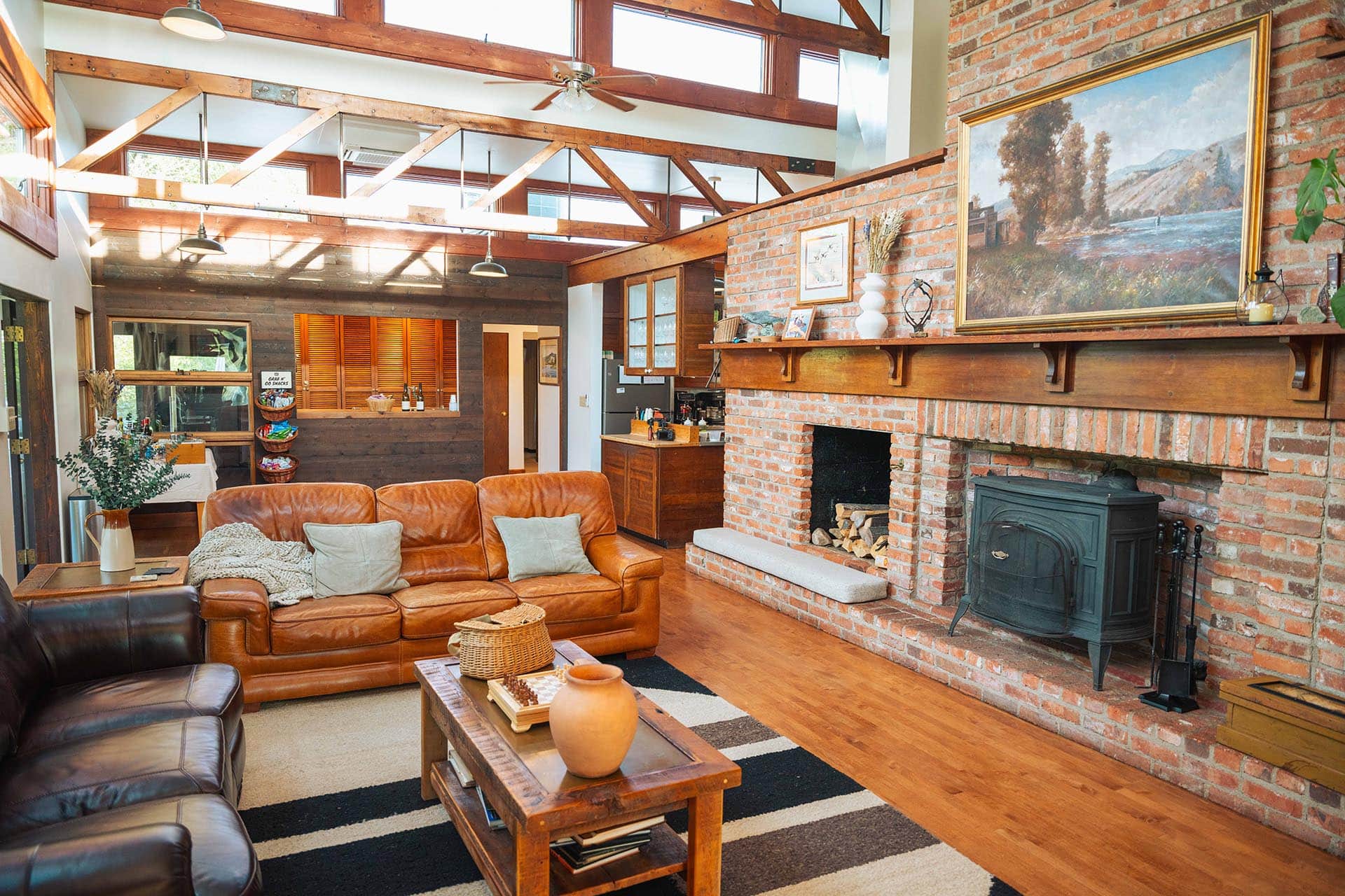Cozy living room with leather sofas, exposed brick fireplace, and wooden ceiling beams in a sunlit open space.