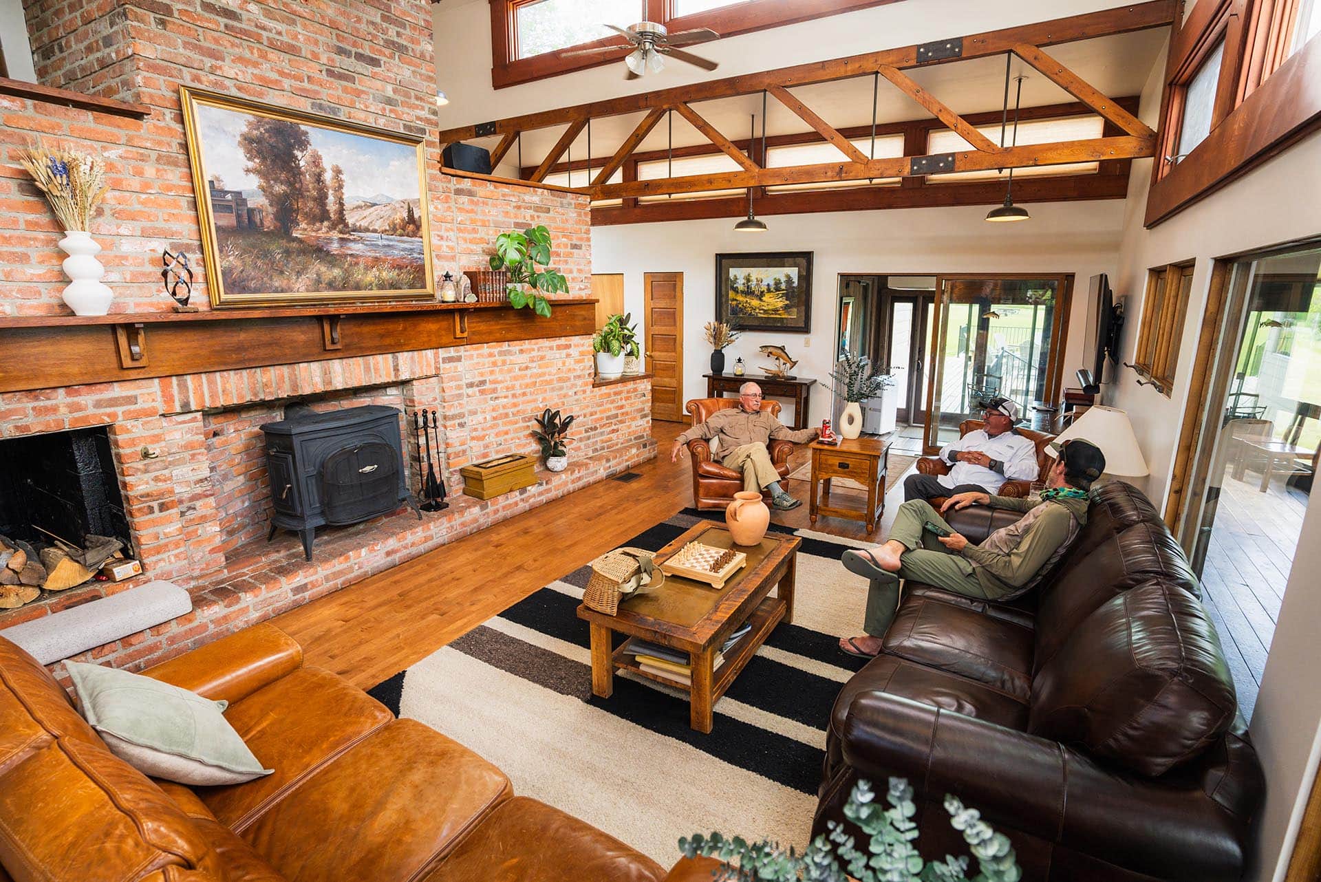 Cozy living room with a brick fireplace, wooden beams, and three men relaxing on brown leather sofas.