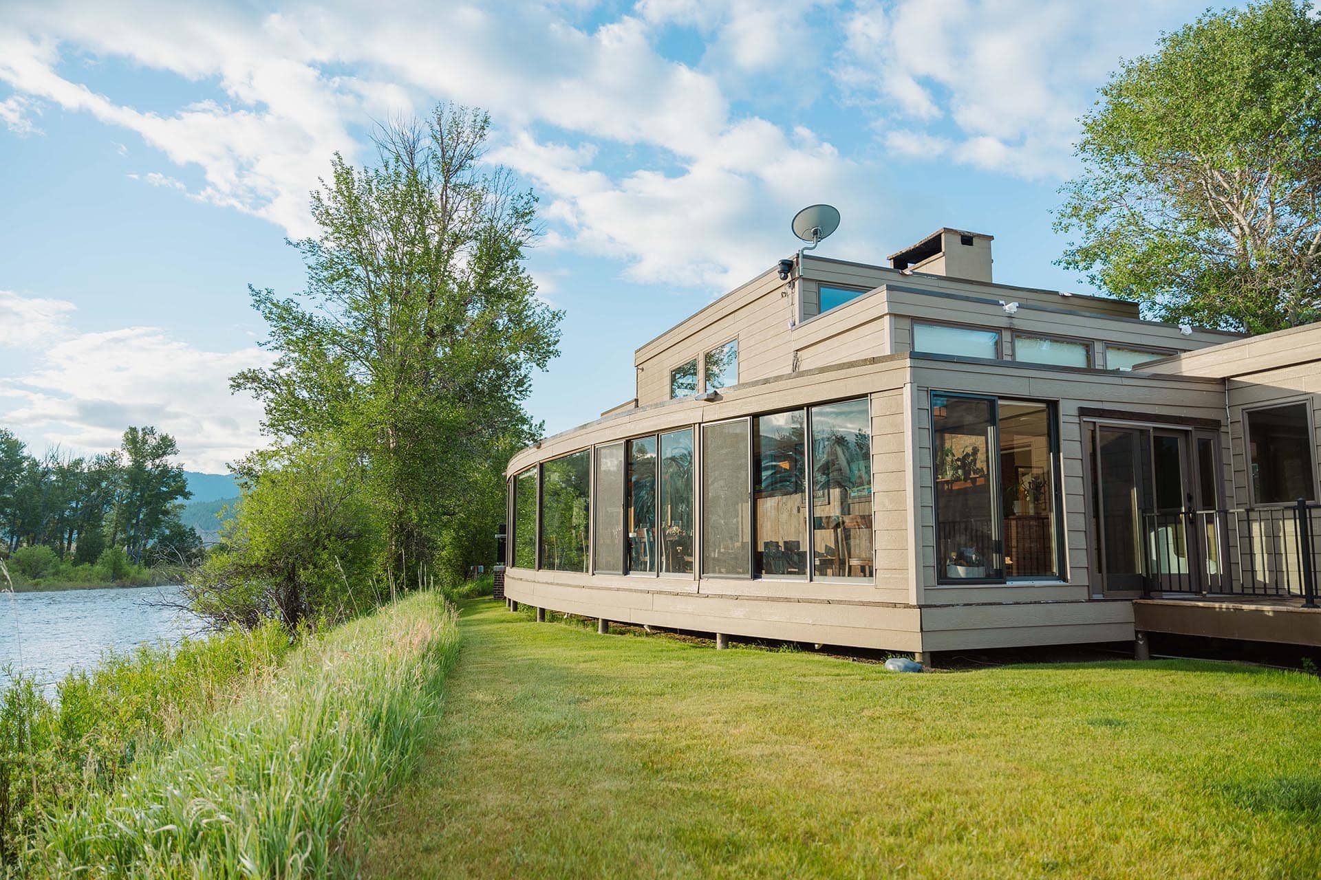 Modern lakeside house with large curved glass walls and a wooden deck, set in green grass and trees under a blue sky.
