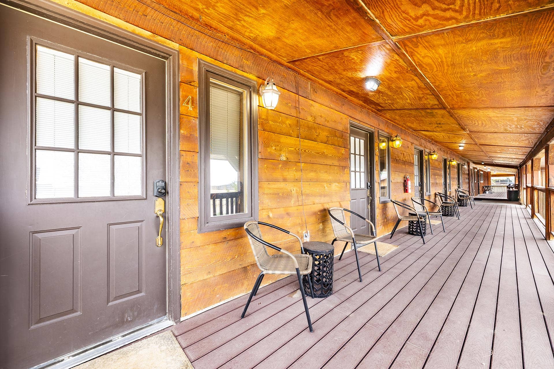 Covered wooden porch with a row of doors and windows, and wicker chairs along the railing under warm lights.