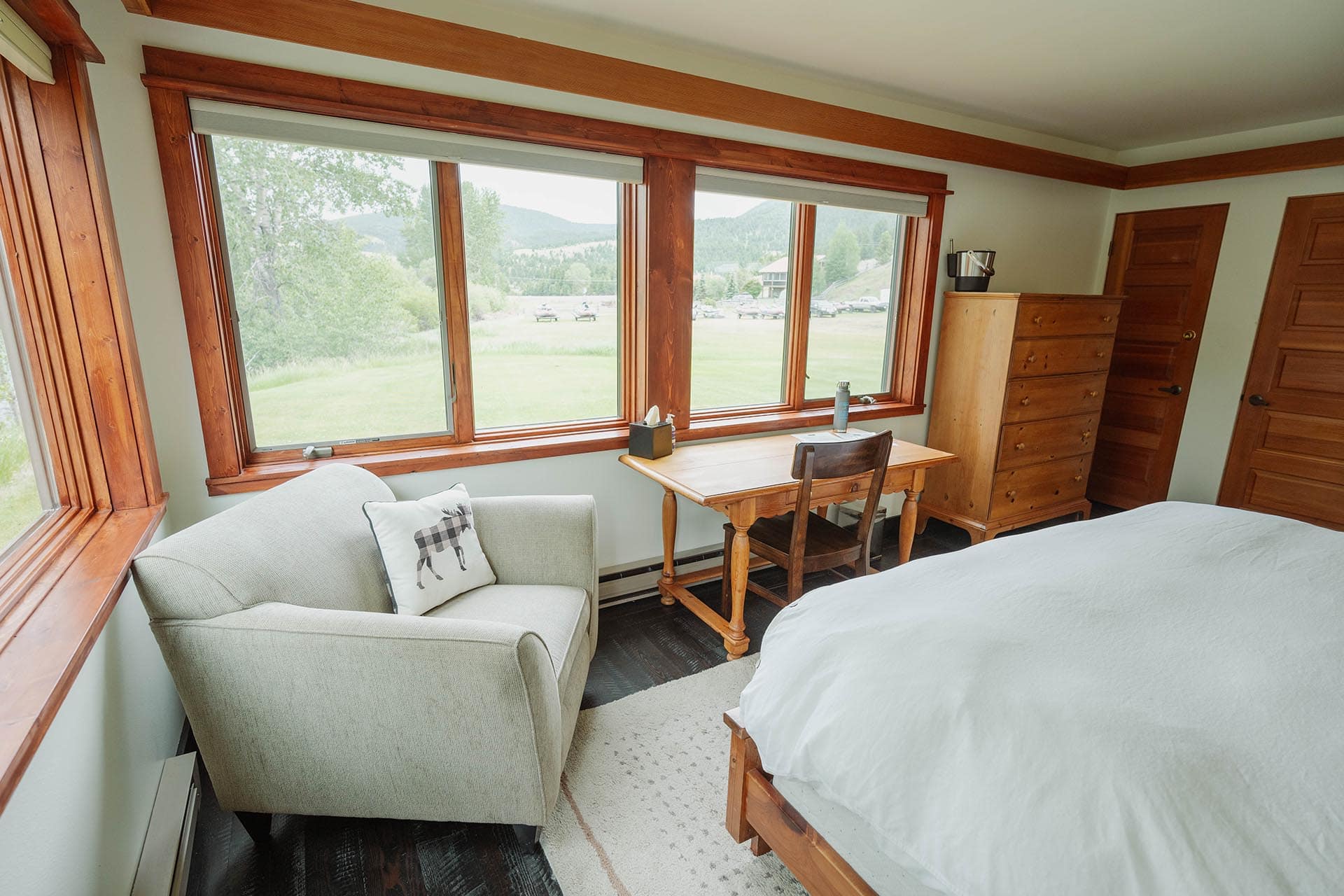 Bedroom with large wooden-framed windows looking out to green lawn and hills; a wooden desk and chair by the window, and a tall wooden dresser inside the room.
