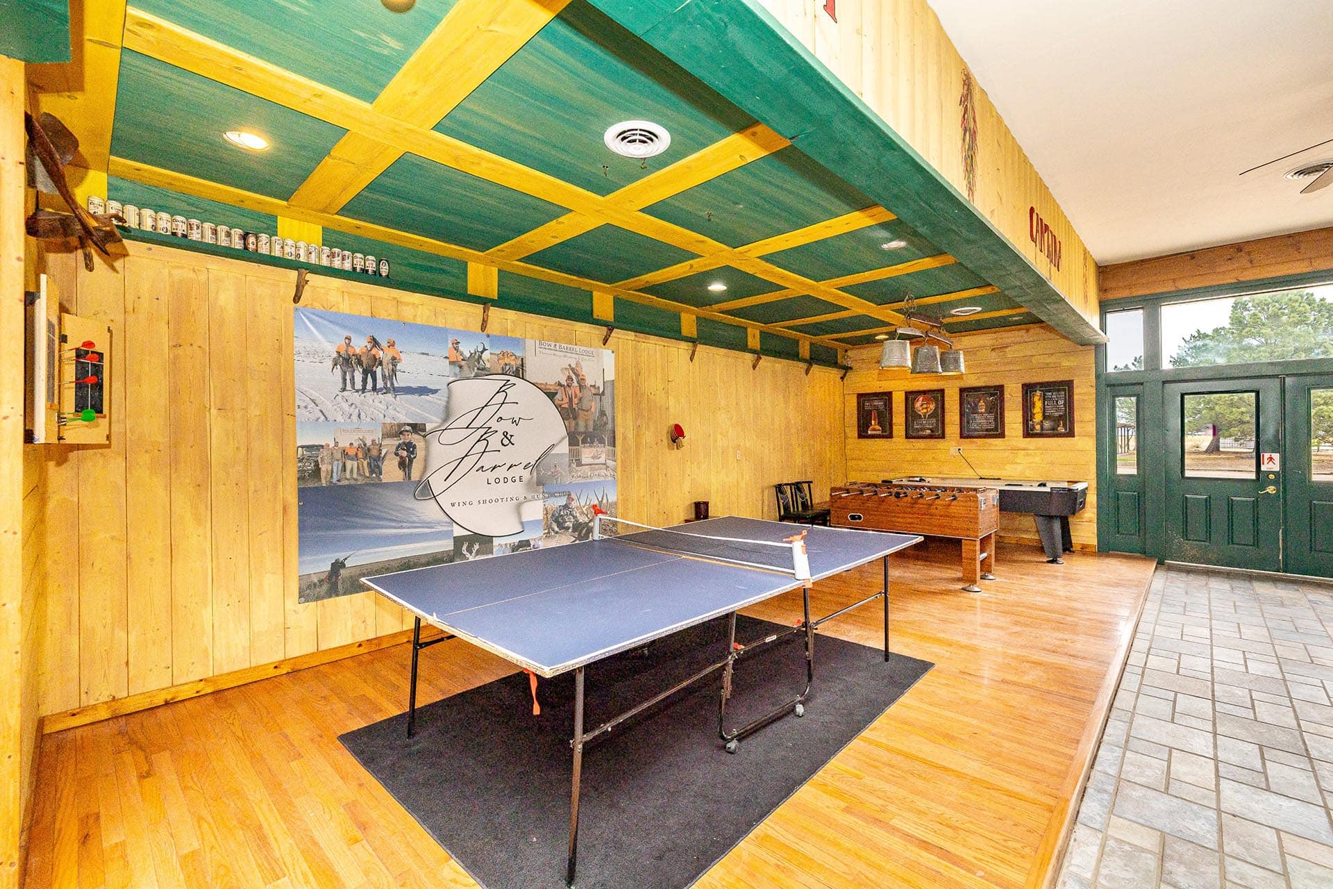 Recreation room in a lodge with a ping-pong table, foosball, wooden walls, and a green ceiling with yellow beams.