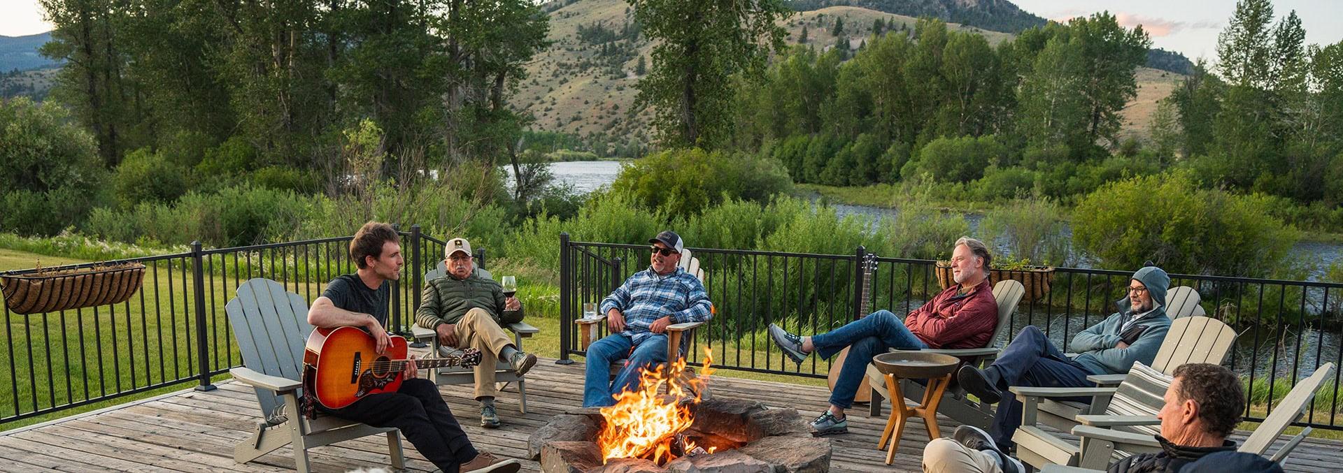 Group of men relaxing on a wooden deck around a campfire; one man plays guitar while others chat.