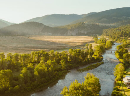 Wide river winds through a sunlit valley with trees and distant mountains on the horizon, a rural settlement to the right.