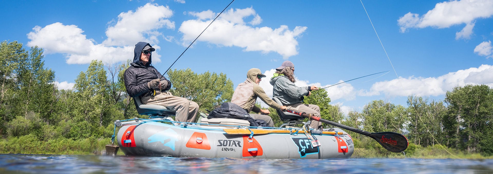 Three people fishing from a large inflatable boat on a calm river, wearing hooded jackets under a bright blue sky with scattered clouds, one cast with a rod into the water.