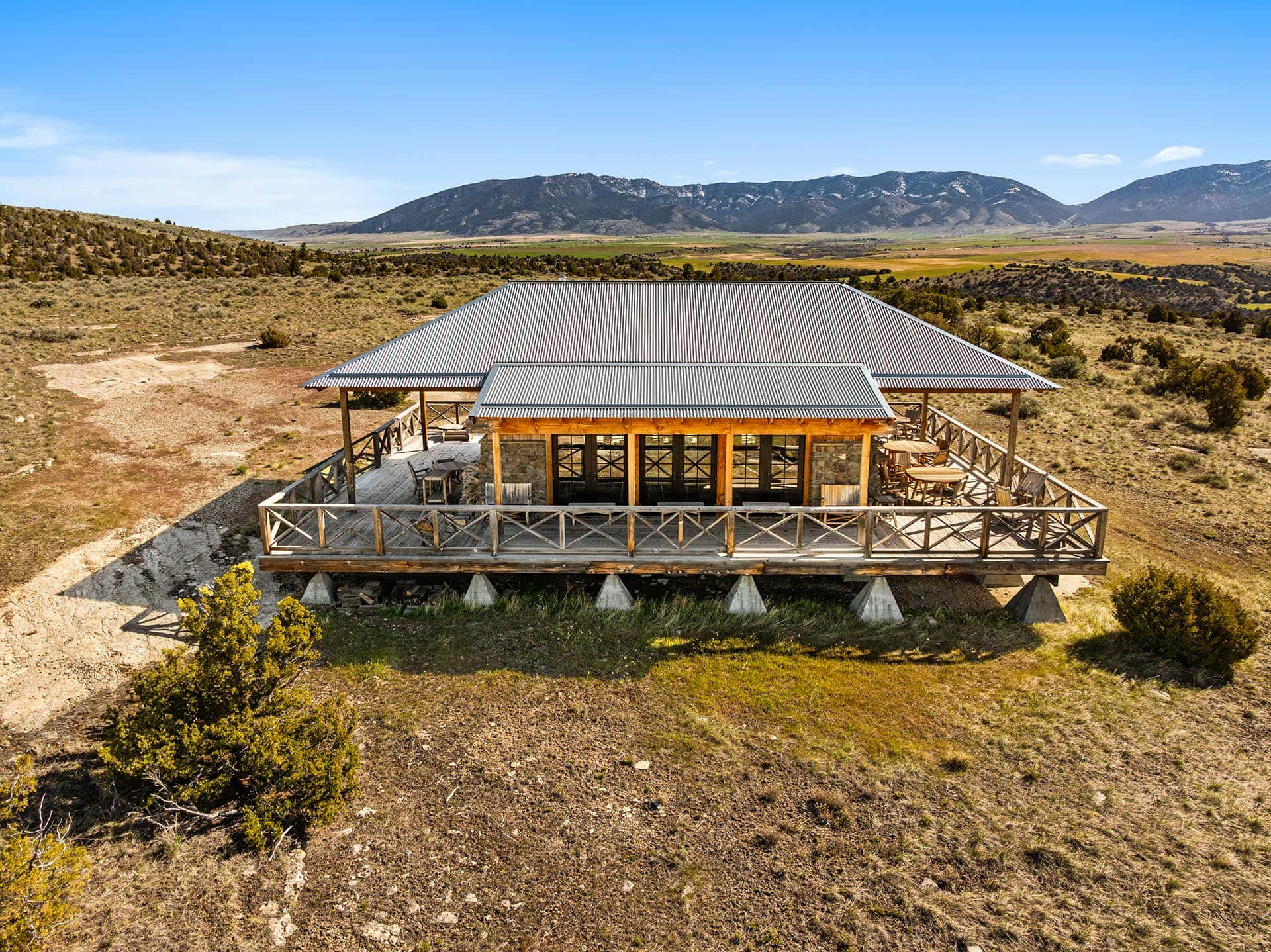 A wooden, covered deck structure with a corrugated metal roof sits on stilts in a dry, scrubby landscape with distant hills and mountains under a bright blue sky.