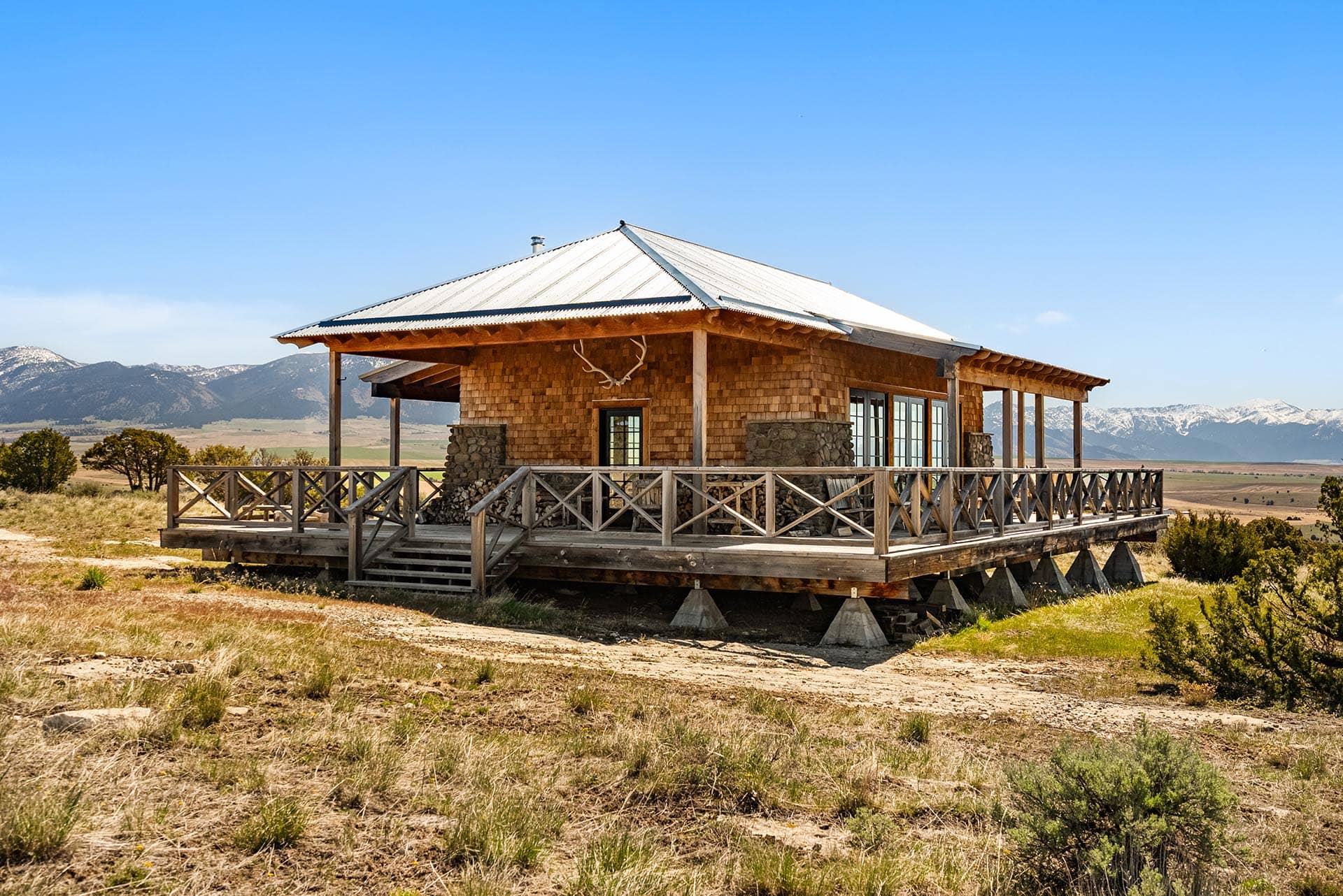 Rustic wooden cabin with a wraparound deck and stone supports set in a dry grassy plain with distant snow-capped mountains under a clear blue sky in the background.