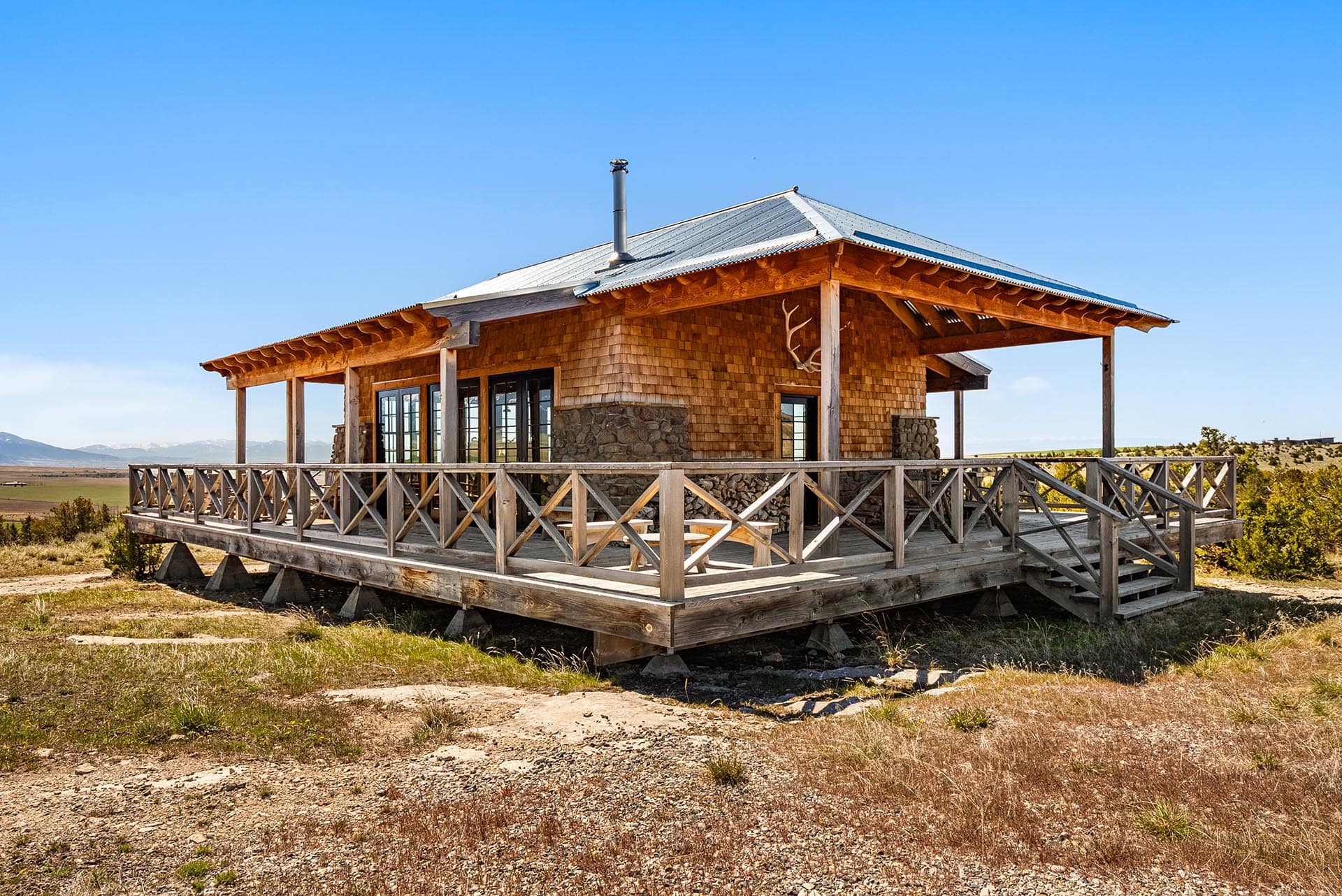 Rustic wooden cabin on a raised deck in a dry field, blue sky and distant mountains visible behind it.
