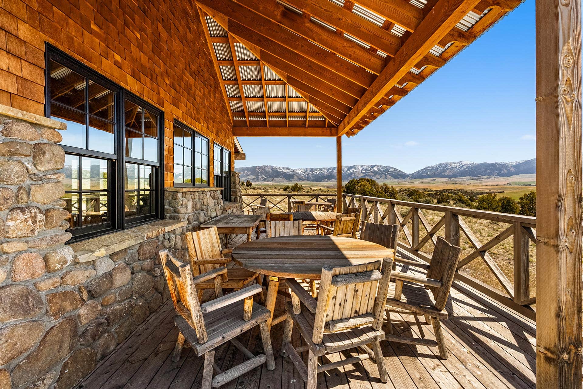 Rustic lodge deck with wooden round tables and chairs, stone wall, overlooking rolling plains and distant mountains.