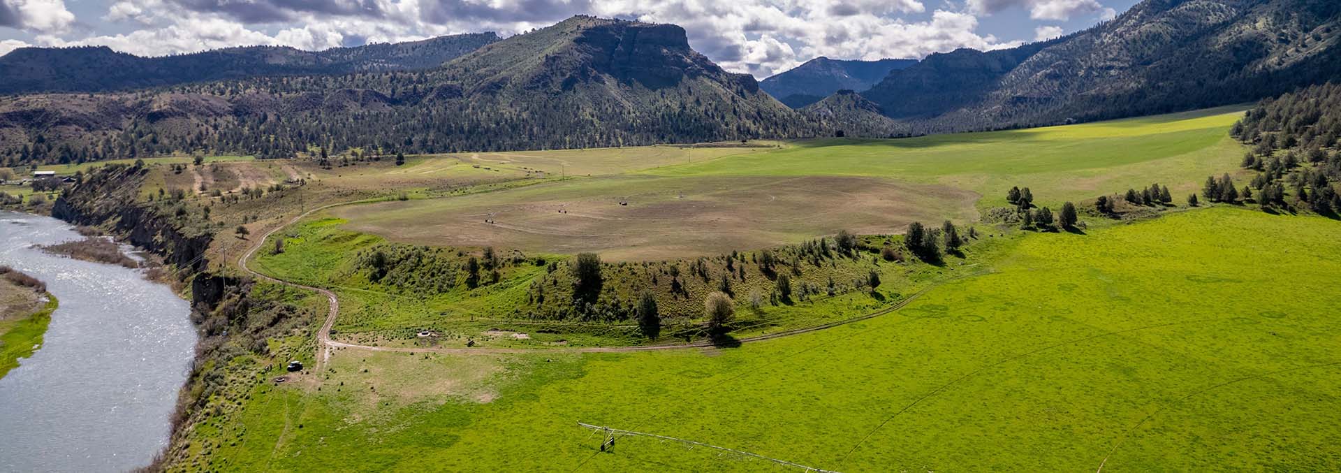 A wide river winds through a green valley with fields, trees, and distant mountains under a partly cloudy sky, aerial view.