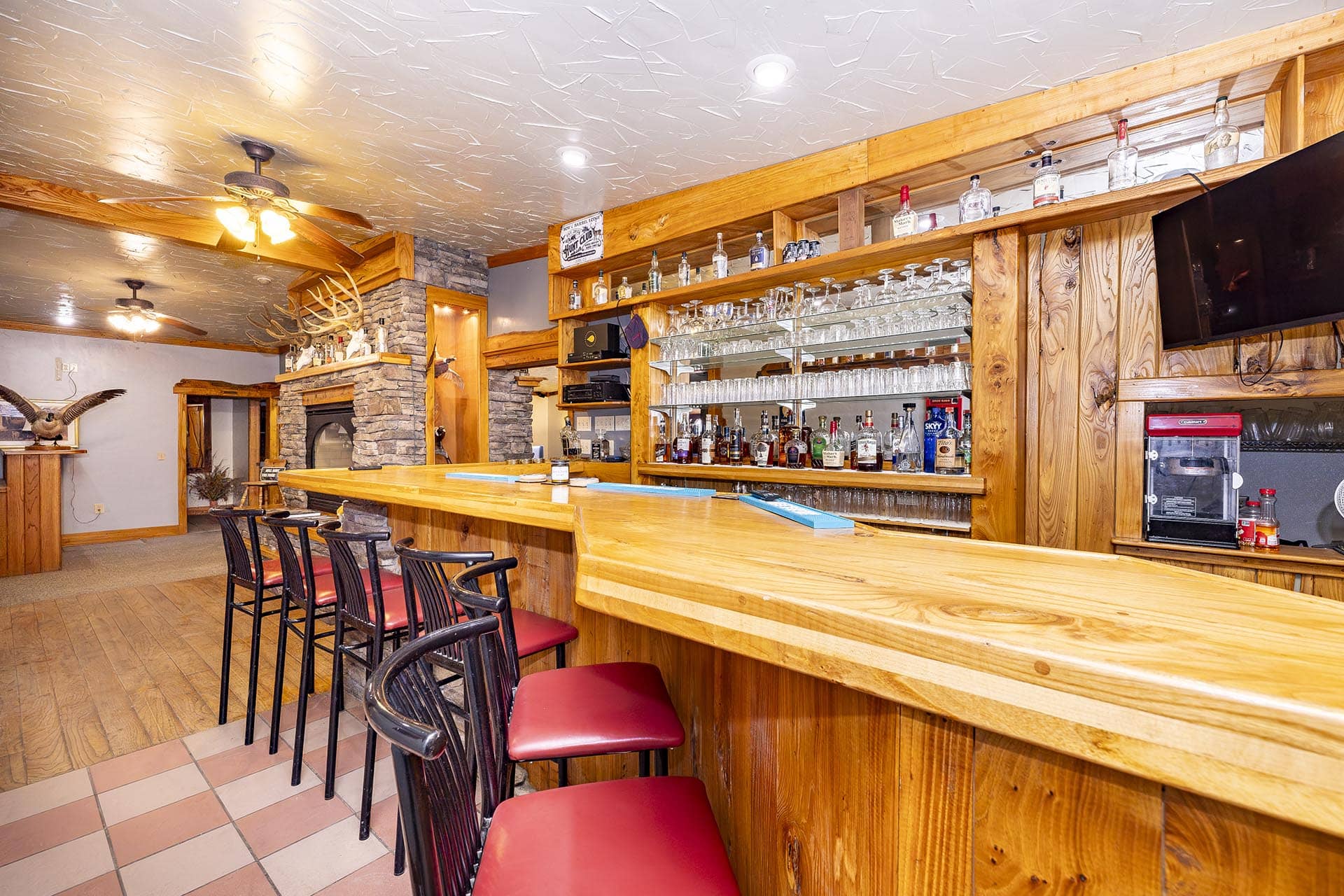 Wooden bar with red-cushioned stools, glassware shelves, and bottles behind it in a rustic tavern setup.