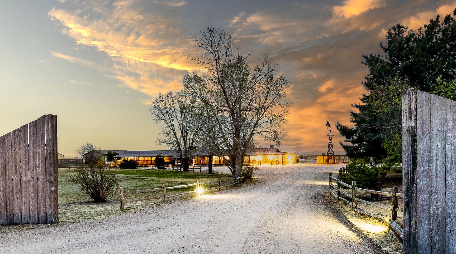 Sunset over a rural farm with a gravel path, wooden gates, and a warmly lit barn in the distance.