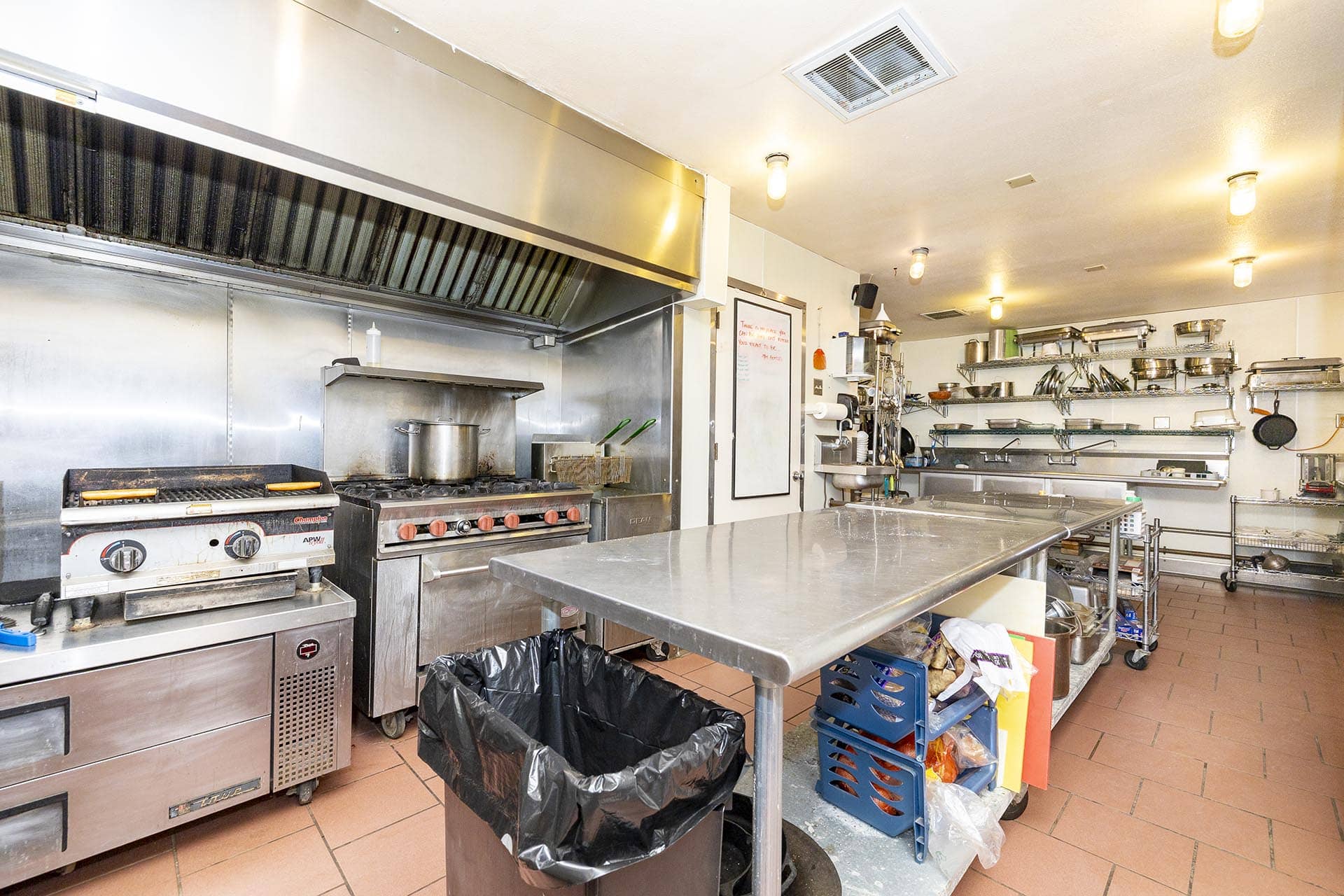 Commercial kitchen with stainless steel appliances, gas range, and overhead hood; cluttered prep table area in view.